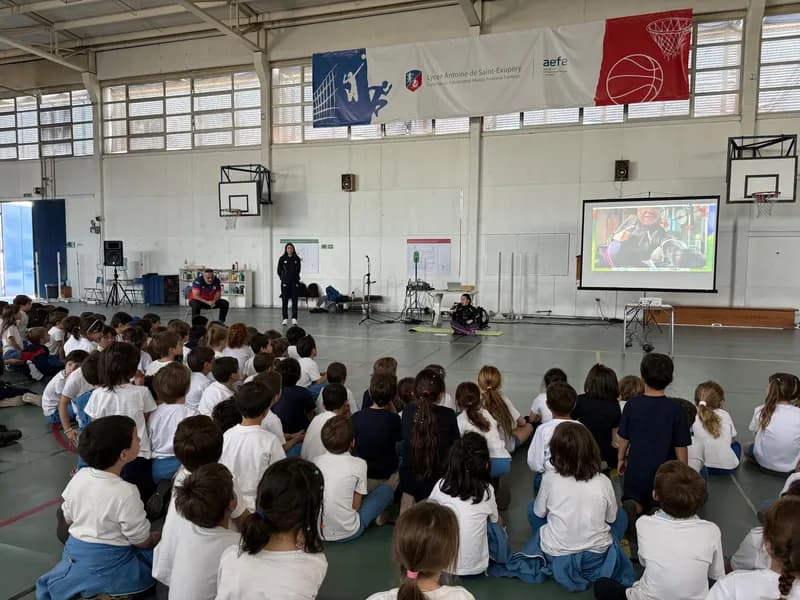 El gimnasio desde el fondo: decenas de estudiantes sentados en el suelo frente a la pantalla, bajo el lienzo del colegio.