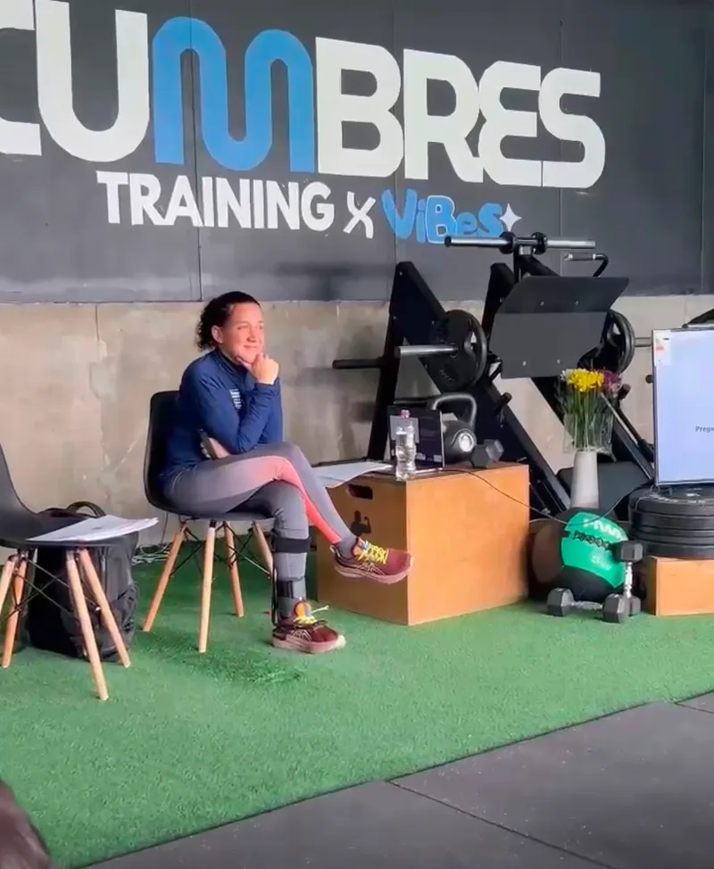 Macarena Larraín sentada frente al grupo, con la mano en el mentón, escuchando. Se le ve la órtesis en la pierna izquierda y detrás el letrero «Cumbres Training».