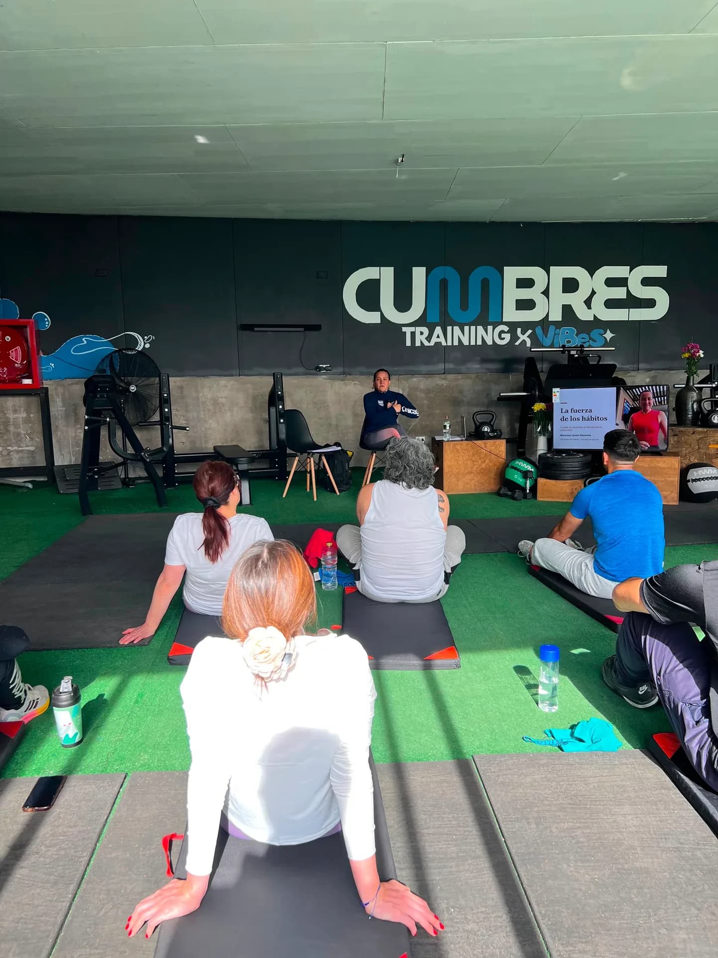La sala de Cumbres Training durante la charla: los asistentes sentados en colchonetas sobre el pasto sintético, y Macarena Larraín al fondo, bajo el letrero del gimnasio, junto a la pantalla que proyecta «La fuerza de los hábitos».