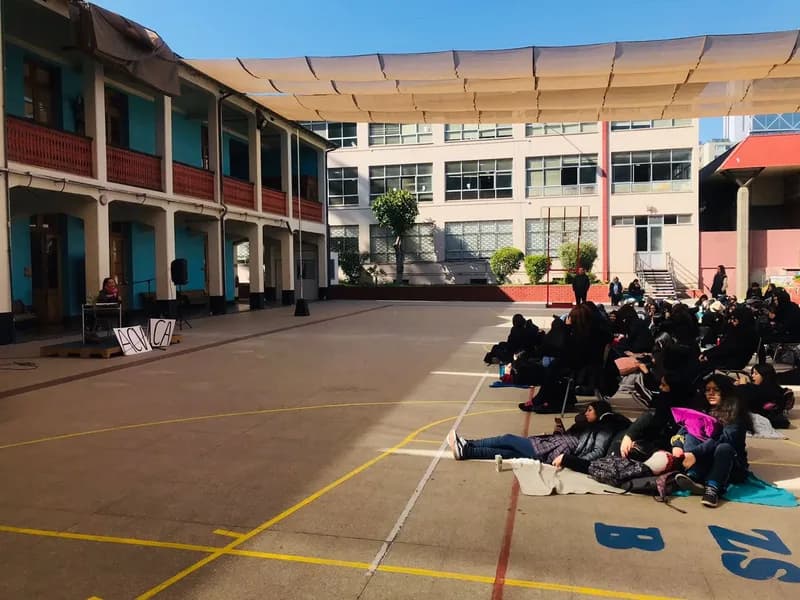 El patio del liceo bajo un toldo: Macarena Larraín habla desde la tarima, a la izquierda, y las estudiantes escuchan sentadas en el suelo, a la derecha.