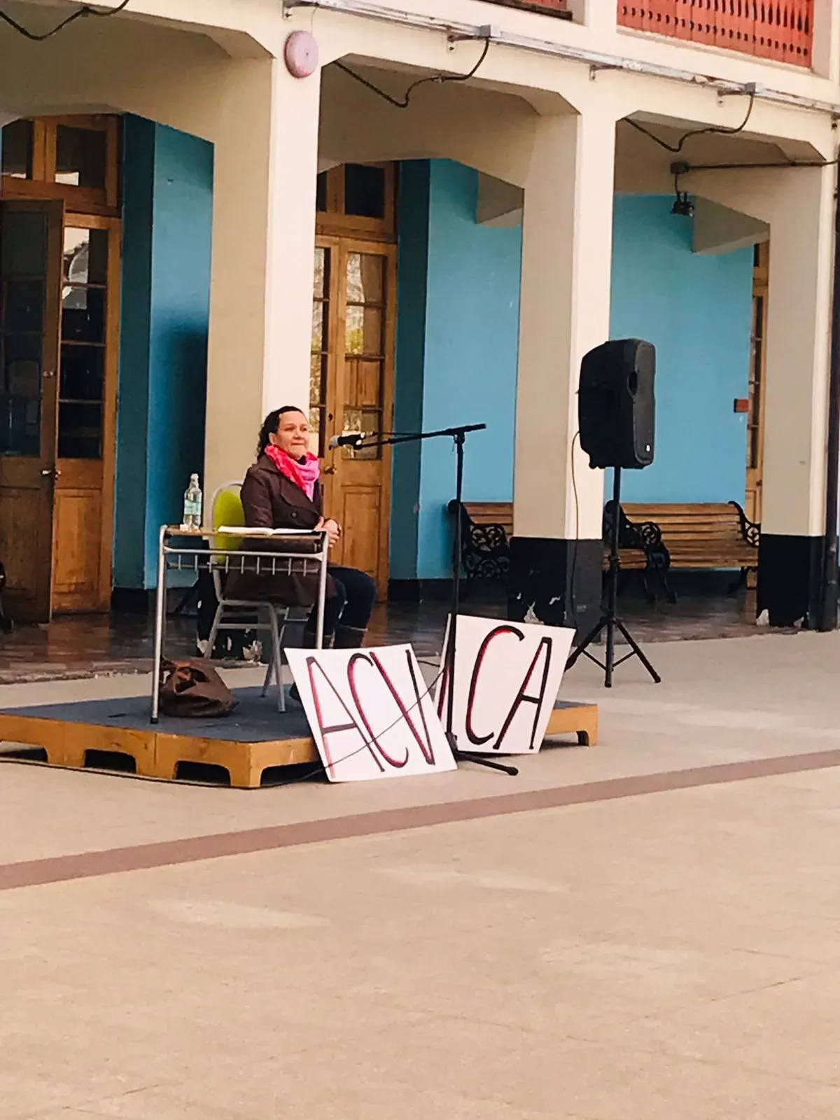 Macarena Larraín, sentada en una tarima del patio del liceo con un micrófono, junto a dos carteles hechos a mano que dicen «ACV».