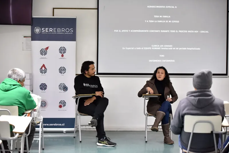 Macarena Larraín conversa con el moderador de SEREBROS, los dos sentados frente a la pantalla que proyecta los agradecimientos del documental.