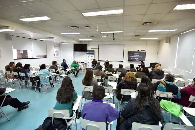 La sala de la Facultad de Psicología llena de asistentes sentados en pupitres, mirando hacia la pantalla y hacia Macarena Larraín al fondo.